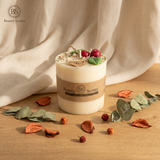 Medium soy candle with cherry wax melt decoration on a wooden table, styled with eucalyptus leaves and dried petals, with soft beige textile backdrop.