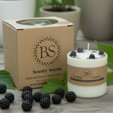 White candle with purple flowers and a brown box labeled 'BS beauty scents' on a wooden surface with blackberries and green leaves.