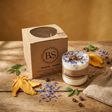 Handmade candle decorated with wildflowers, next to branded packaging, coffee beans, and leaves on a warm-toned wooden tabletop.
