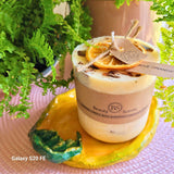 Handmade scented candle by Beauty Scents, decorated with dried orange slices and cinnamon sticks, placed on a yellow ceramic dish surrounded by green ferns.