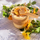 Decorative candle with orange slices and a tag, surrounded by flowers on a textured surface.