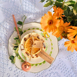 Handmade scented candle by Beauty Scents, topped with dried orange slices and cinnamon sticks, displayed on a floral plate with bright orange flowers and a copper candle snuffer on a lace tablecloth.