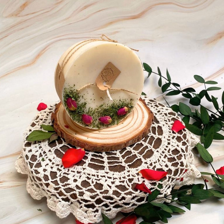 Decorative candle in a wooden holder on a lace doily with red flowers and green leaves.