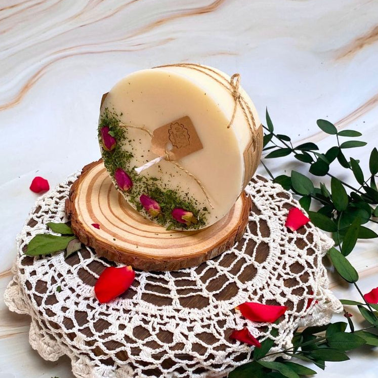 Candle in a wooden holder on a lace doily with greenery and rose petals.