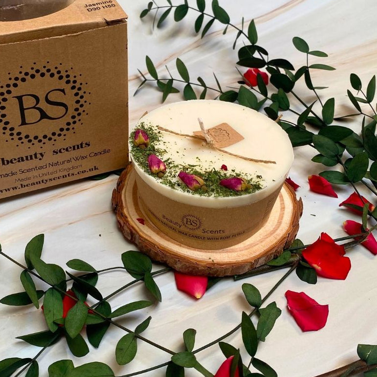 Decorative candle with rose petals on a wooden coaster, surrounded by greenery and red flowers, with Beauty Scents branding.