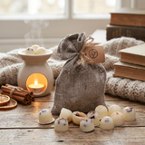 Grey linen bag filled with handmade orange and cinnamon scented soy wax melts with shredded cinnamon, displayed on a rustic wooden table next to an oil burner, cinnamon sticks, and dried orange slices.
