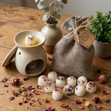Grey linen bag filled with handmade soy wax melts decorated with rose petals, displayed on a rustic wooden table with an oil burner, dried flowers, and green plants.