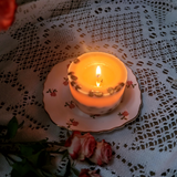 Burning small coffee candle with warm golden flame on vintage pink floral plate surrounded by pink roses and white lace doily creating romantic ambiance