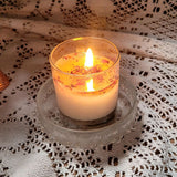 Small glass rose petals scented candle with natural botanicals burning on vintage white lace, showing warm amber flame and melted wax pool