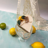 Candle with a tag on a glass stand surrounded by lemons and limes on a textured surface.