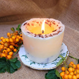 Large handmade scented candle by Beauty Scents, burning with a bright flame, topped with shredded cinnamon sticks, displayed on a vintage plate with orange berries and green leaves, set against a rustic burlap background.