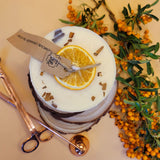 Large handmade scented candle by Beauty Scents, topped with a dried orange slice and cinnamon stick pieces, displayed on rustic wooden coasters with orange berries and copper candle tools.