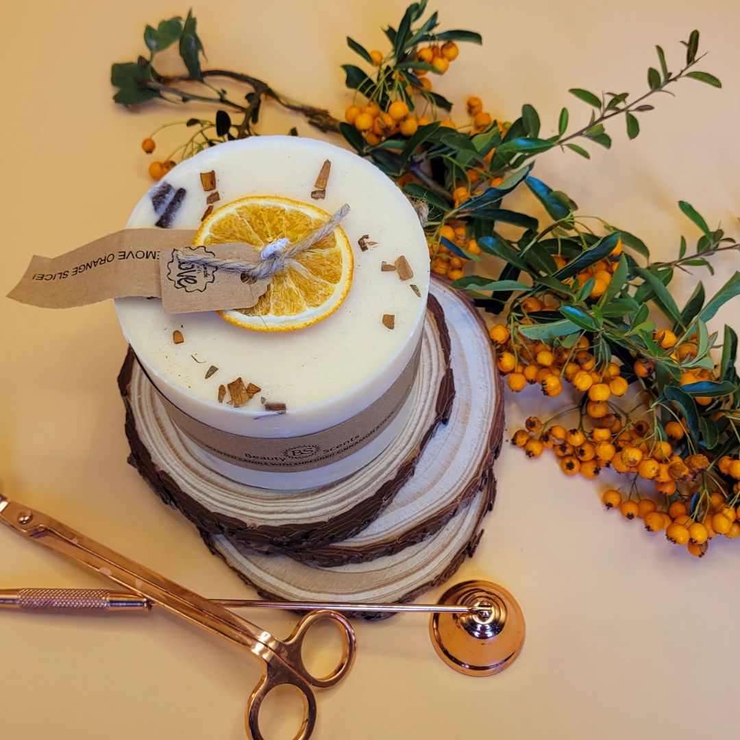 Large handmade scented candle by Beauty Scents, decorated with a dried orange slice and cinnamon sticks, displayed on rustic wood slices with orange berries and copper candle tools on a beige background.