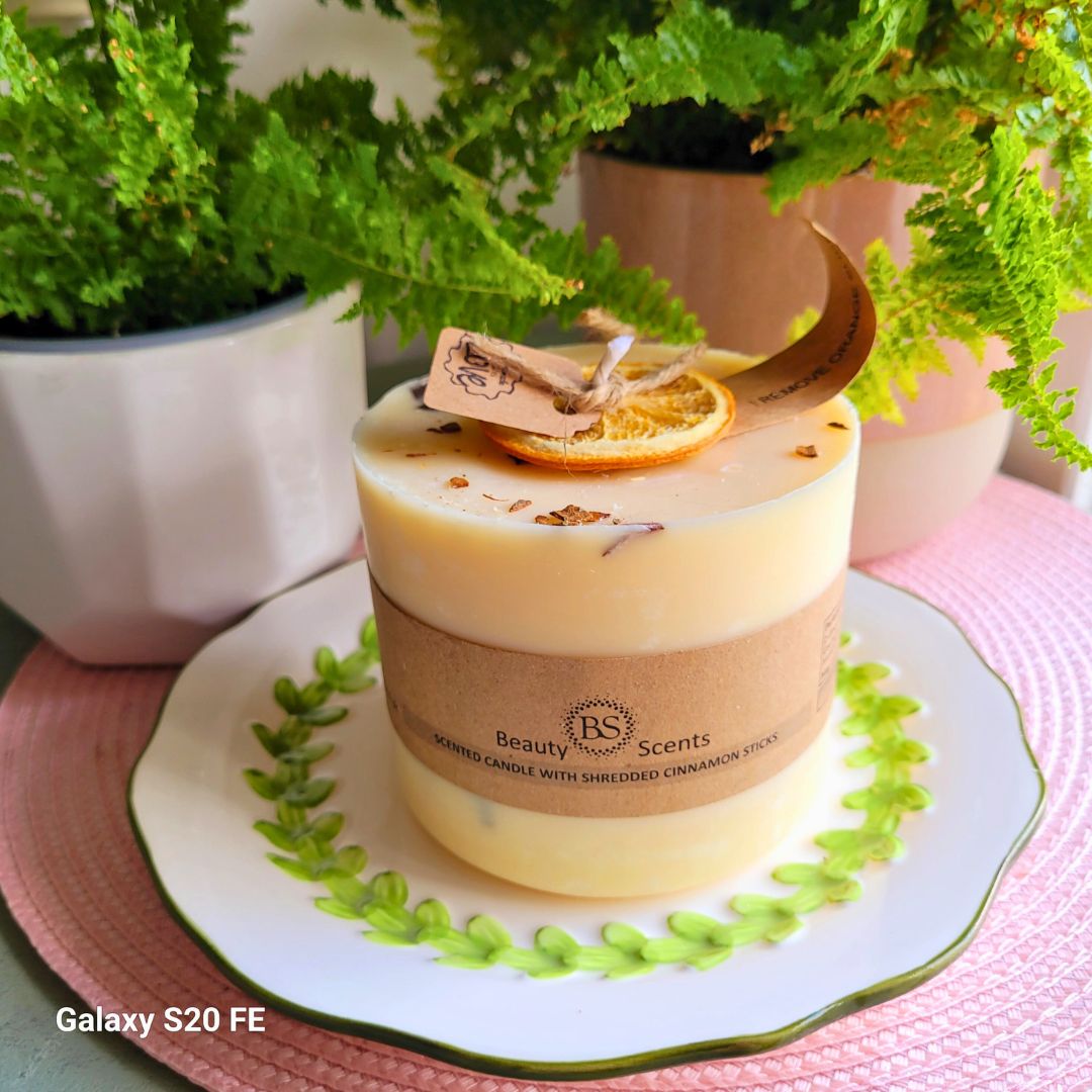 Large handmade scented candle by Beauty Scents, decorated with dried orange slice and cinnamon sticks, placed on a white plate with green leaf design, surrounded by potted ferns.