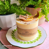 Large handmade scented candle by Beauty Scents, decorated with dried orange slice and cinnamon sticks, placed on a white plate with green leaf design, surrounded by potted ferns.