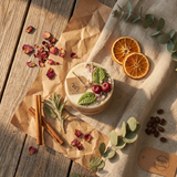 Small soy candle with cherry wax melt decoration and green leaf detail on white textile, styled with dried orange slices, eucalyptus leaves, cinnamon sticks and scattered dried petals.