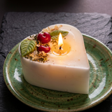Close-up of a lit heart-shaped soy candle with a melted wax pool, decorated with cherry wax melts and green leaf details on a green ceramic plate.
