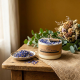 Beauty Scents small candle with blue petals and kraft label, next to a dish of petals and natural bouquet on wood.