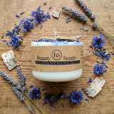 Handmade candle with wildflowers, lying sideways on wood, styled with blue cornflower petals and rustic natural elements.