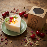 Lit heart-shaped soy candle with cherry wax melt decoration on a green ceramic plate, styled on a rustic wooden table next to a Beauty Scents gift box with fresh cherries and dried petals.