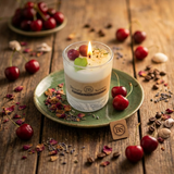 Lit small soy candle in a glass jar with wax cherries and green leaf decoration on a green ceramic plate on a rustic wooden table, styled with fresh cherries and dried petals.
