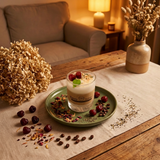 Small soy candle in a glass jar with wax cherries and green leaf decoration on a green plate on a wooden table in a cosy living room at night, styled with dried petals and coffee beans.