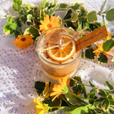 Small handmade scented candle by Beauty Scents in a glass jar, topped with a dried orange slice and cinnamon sticks, surrounded by yellow flowers and green leaves on a white lace cloth.
