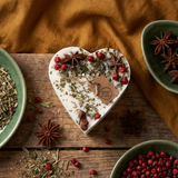 Heart-shaped candle decorated with herbs and spices on a wooden surface with bowls of additional spices
