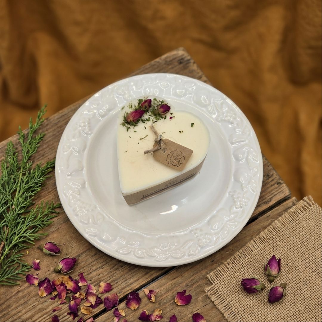 Heart-shaped candle on a white decorative plate with dried flowers and a tag on a wooden surface.