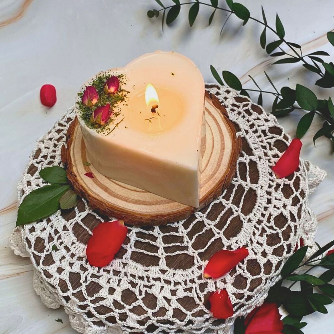 Heart-shaped candle on a wooden block with a slice cut out, surrounded by red flowers and green leaves on a white background.