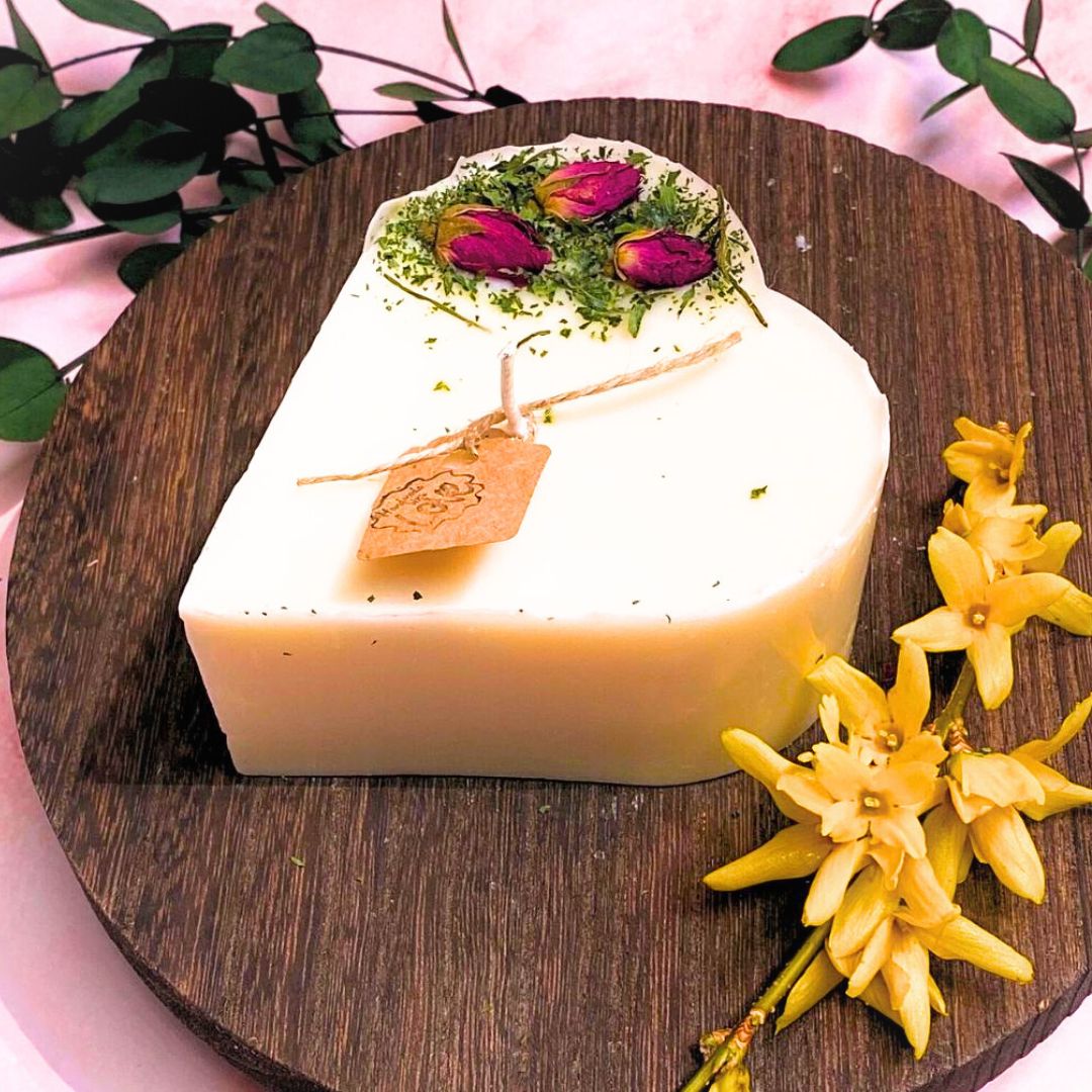 Small heart-shaped soy candle decorated with real rose buds and greenery, handmade by Beauty Scents, displayed on a wooden board with yellow flowers