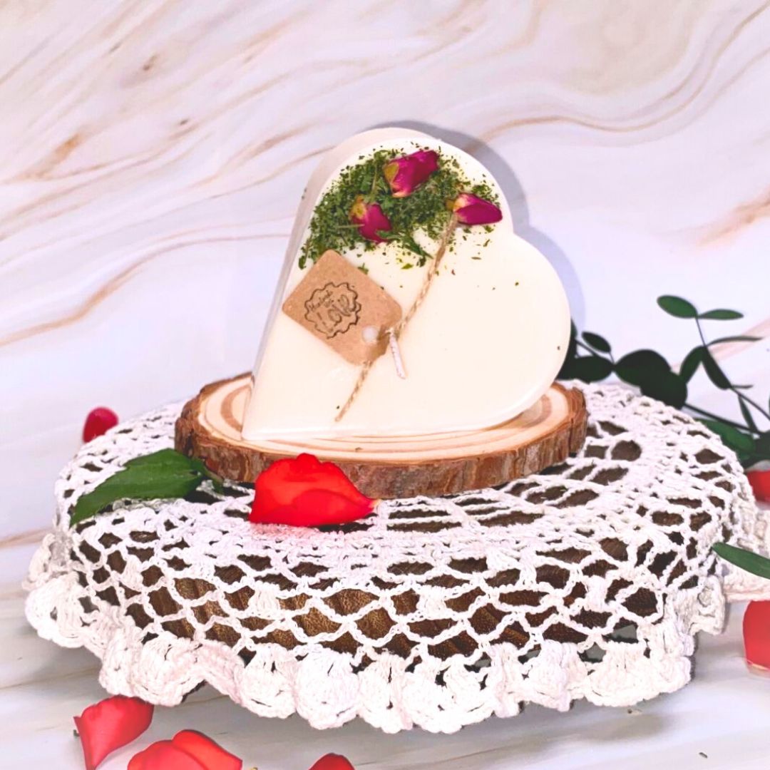 Small heart-shaped soy candle decorated with real rose buds and greenery, handmade by Beauty Scents, displayed on a wooden board with yellow flowers