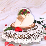 Small heart-shaped soy candle decorated with real rose buds and greenery, handmade by Beauty Scents, displayed on a wooden board with yellow flowers