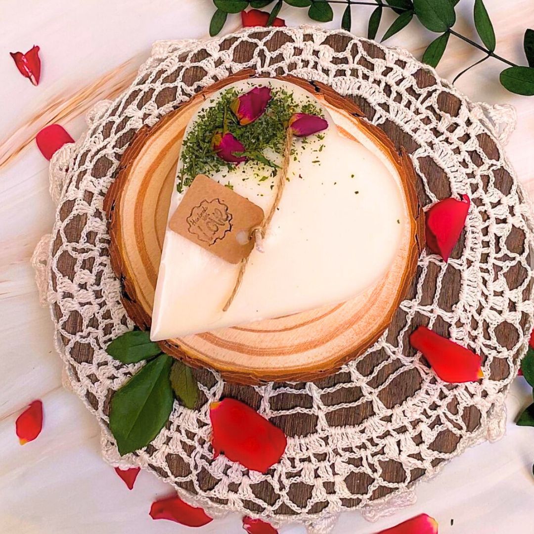 Small heart-shaped soy candle topped with real rose buds and greenery, handmade by Beauty Scents, styled on a wooden slice with lace and scattered rose petals