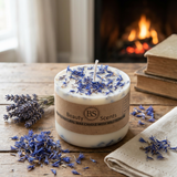 Small Beauty Scents wildflower candle topped with blue cornflower petals, displayed on a rustic wooden table with dried lavender, books, and a fireplace in the background, square image.