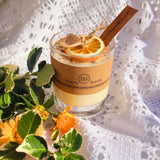 Small handmade scented candle by Beauty Scents in a glass jar, decorated with a dried orange slice and cinnamon sticks, surrounded by yellow flowers and green leaves on white lace fabric.