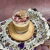 Handmade rose petal candle in glass jar displayed on wooden platform with dried botanicals and white crochet lace