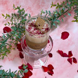 Small glass rose petals soy candle with Beauty Scents label displayed on glass stand with eucalyptus and scattered rose petals