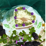 Decorative candle with purple flowers and green leaves on a white plate with gold rim, surrounded by blackberries and green leaves.