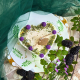 Heart-shaped candle with greenery and purple flowers on a decorative plate with green leaves and blackberries.