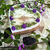 Large heartsheped candle with purple flowers and green leaves on a textured surface with blackberries and greenery.