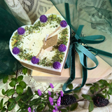 Heart-shaped gift box with greenery and purple flowers on a textured surface with greenery.