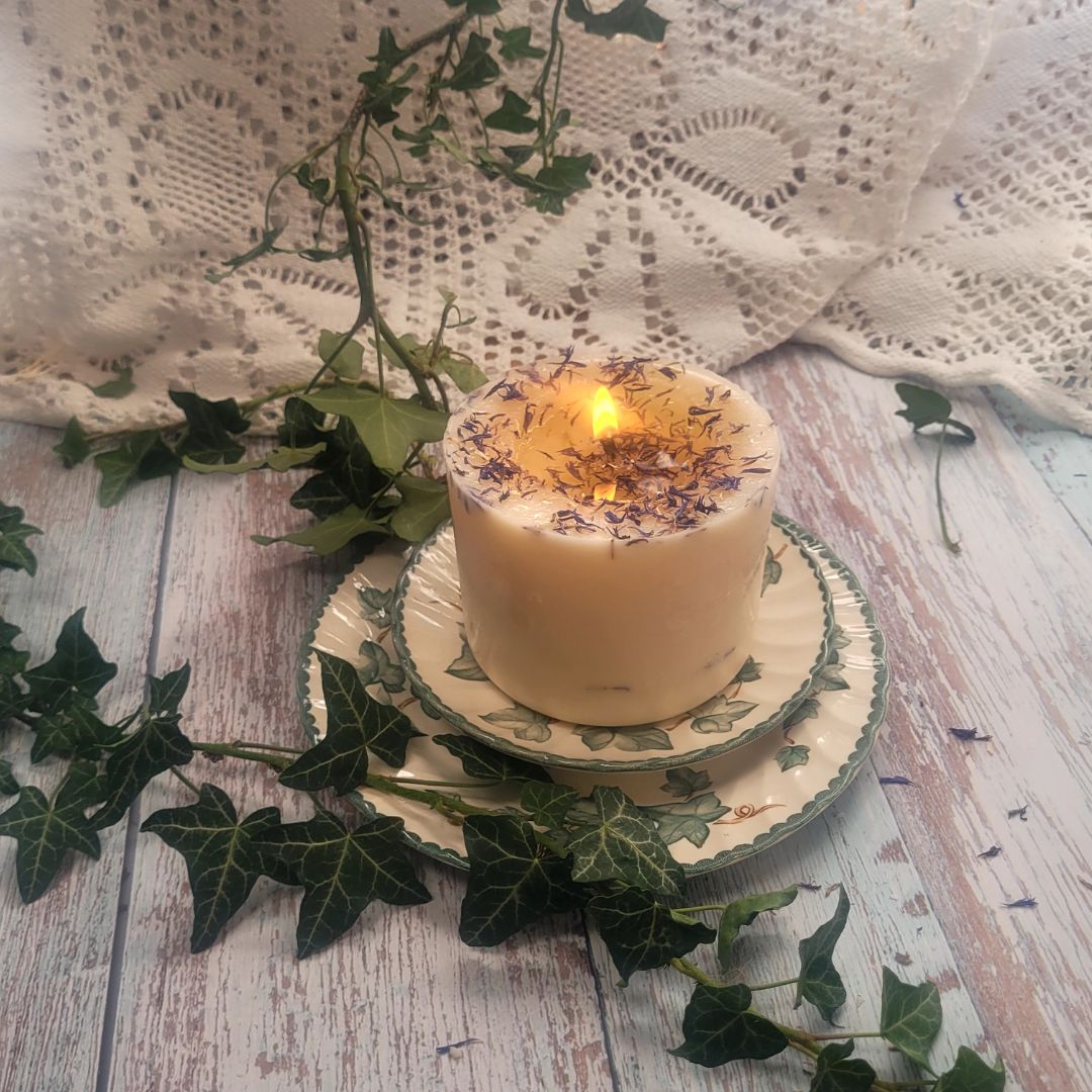 Large Wild Flowers soy candle 700g burning with lit flame and melted wax pool, blue cornflowers decoration visible, styled on ivy-patterned plate with fresh ivy and white lace - candle burning demonstration photography
