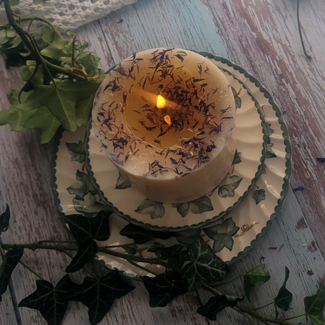 Large Wild Flowers soy candle 700g burning top-down view with lit flame and wax pool, blue cornflowers decoration around edges, styled on ivy-patterned plate with fresh ivy and lace - overhead burning demonstration photography