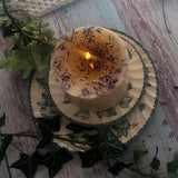 Large Wild Flowers soy candle 700g burning top-down view with lit flame and wax pool, blue cornflowers decoration around edges, styled on ivy-patterned plate with fresh ivy and lace - overhead burning demonstration photography