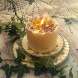Large Wild Flowers soy candle 700g burning with lit flame and melted wax pool, blue cornflowers decoration visible, styled on ivy-patterned plate with fresh ivy and white lace - candle burning demonstration photography