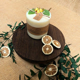 Candle with decorative elements on a wooden board surrounded by greenery on a textured surface