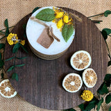Medium lemon candle with botanicals on rustic wooden board with dried citrus slices and yellow flowers on burlap