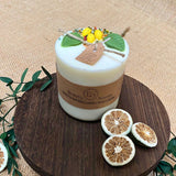 White candle with decorative elements on a wooden board with green leaves and dried citrus slices.
