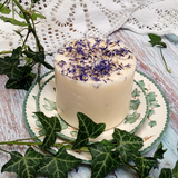 Medium Wild Flowers soy candle 500g with blue cornflowers decoration on white wax, styled with fresh ivy vines on vintage leaf-patterned ceramic plate, rustic whitewashed wood background